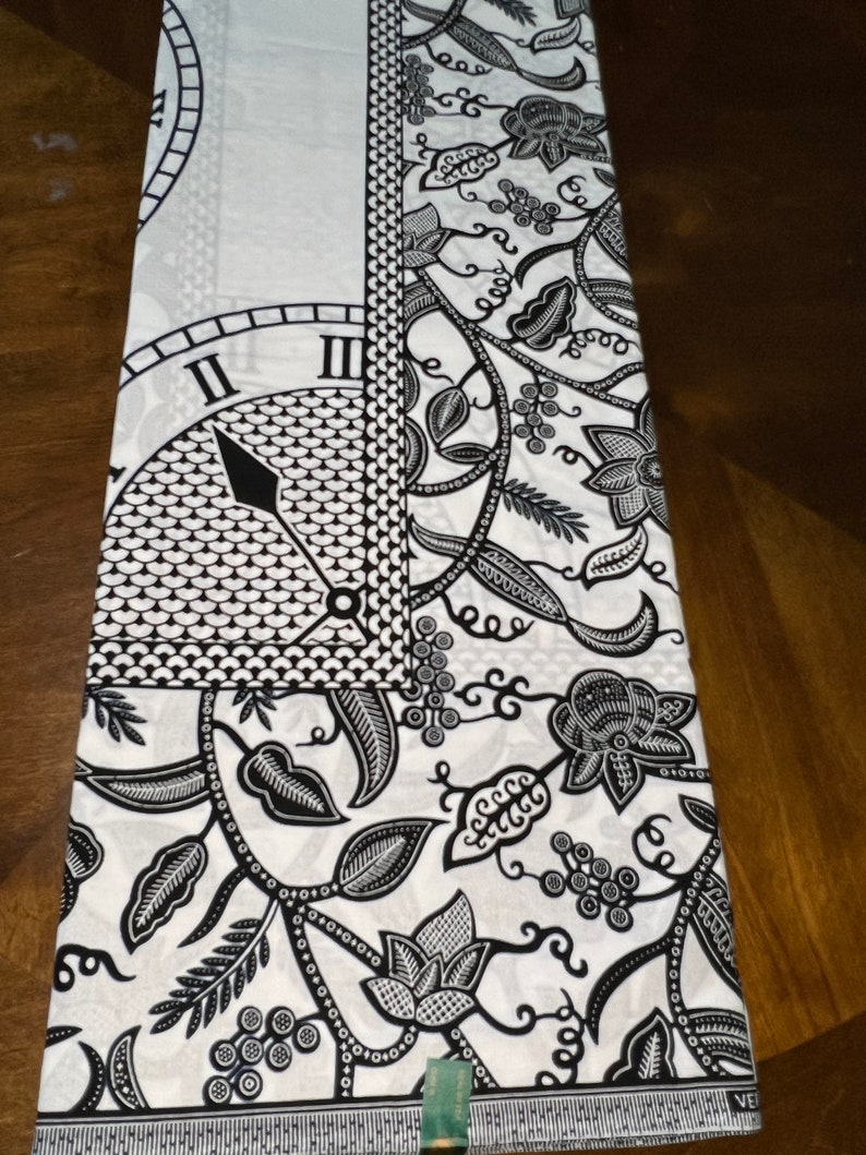 May include: Black and white fabric with a floral pattern and a clock face design. The fabric has a white background with black floral vines, leaves, and flowers. The clock face is in the upper left corner and has black Roman numerals and a black hand.