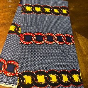 K&ouml;nnte beinhalten: Blauer Stoff mit einem sich wiederholenden Kettenmuster in Rot, Gelb und Schwarz. Der Stoff hat einen kleinen wei&szlig;en und blauen geometrischen Hintergrund. Die W&ouml;rter "SPEED DUTCH WAX" und "VERITABLE W" sind sichtbar.