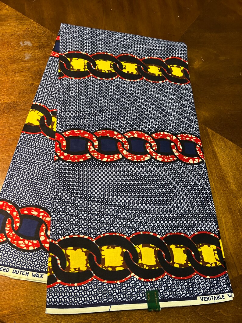 K&ouml;nnte beinhalten: Blauer Stoff mit einem sich wiederholenden Kettenmuster in Rot, Gelb und Schwarz. Der Stoff hat einen kleinen wei&szlig;en und blauen geometrischen Hintergrund. Die W&ouml;rter "SPEED DUTCH WAX" und "VERITABLE W" sind sichtbar.