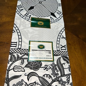 May include: Black and white fabric with a clock and floral pattern. The fabric has a label that says "Guaranteed Real Dutch Wax Block Print" and "Printed in Holland".