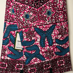 Op de afbeelding: Een gevouwen stof met een levendig patroon. Het ontwerp heeft een roze achtergrond met witte, zwarte en blauwe accenten. De stof heeft geometrische vormen, stervormen en bladmotieven. Het label luidt "PRINTED BY".