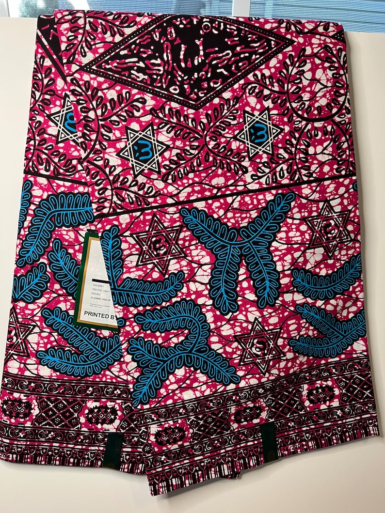 Op de afbeelding: Een gevouwen stof met een levendig patroon. Het ontwerp heeft een roze achtergrond met witte, zwarte en blauwe accenten. De stof heeft geometrische vormen, stervormen en bladmotieven. Het label luidt "PRINTED BY".