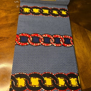 K&ouml;nnte beinhalten: Gerollter Stoff mit einem sich wiederholenden Muster aus kleinen wei&szlig;en und blauen Kreisen. Der Stoff weist B&auml;nder mit kettenartigen Designs in Rot, Gelb und Schwarz auf. Ein gr&uuml;nes Etikett lautet "PRINTED IN HOLLAND". Der Stoff ist mit "VERITABLE W" gekennzeichnet.