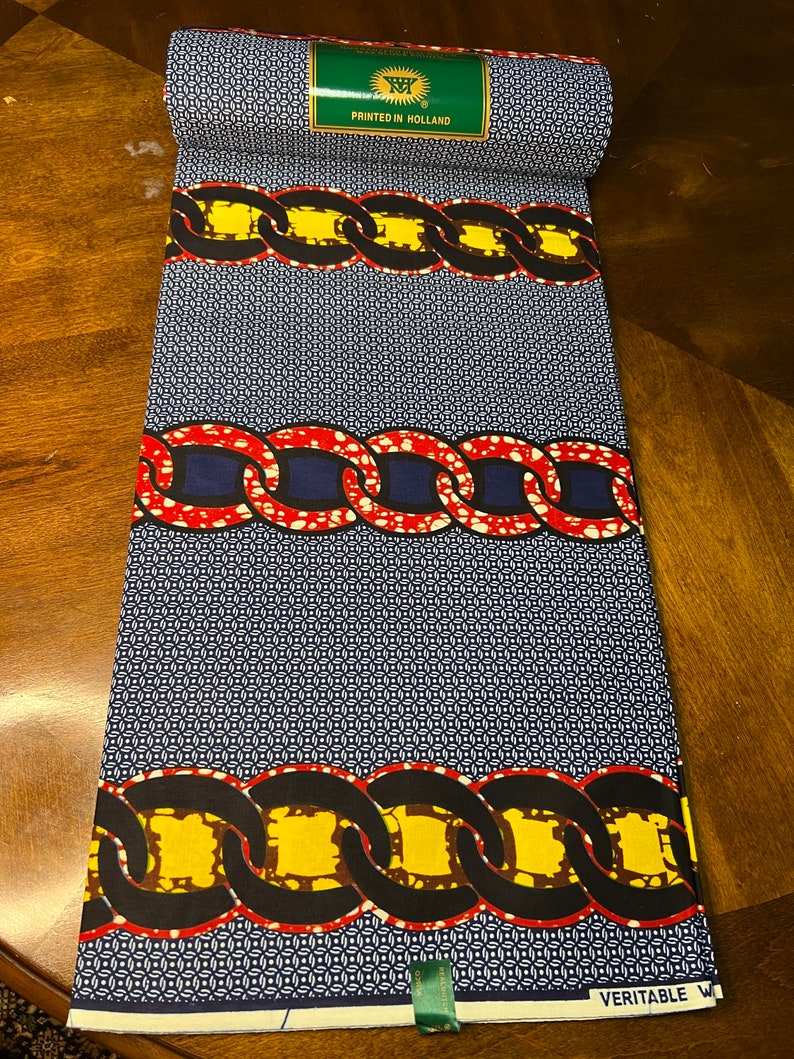 K&ouml;nnte beinhalten: Gerollter Stoff mit einem sich wiederholenden Muster aus kleinen wei&szlig;en und blauen Kreisen. Der Stoff weist B&auml;nder mit kettenartigen Designs in Rot, Gelb und Schwarz auf. Ein gr&uuml;nes Etikett lautet "PRINTED IN HOLLAND". Der Stoff ist mit "VERITABLE W" gekennzeichnet.