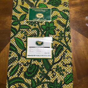 K&ouml;nnte beinhalten: Ein gelber und gr&uuml;ner Stoff mit einem Blumenmuster. Der Stoff ist bedruckt mit dem Text "Guaranteed Real Dutch Wax Block Prints" und "Printed in Holland".