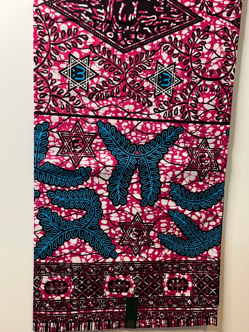 Op de afbeelding: Een levendige stof met een herhalend patroon van roze, zwarte en blauwe ontwerpen. De stof heeft Davidster-vormen, bladachtige motieven en geometrische randen. Het algehele ontwerp is een mix van gedurfde kleuren en ingewikkelde details.