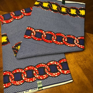 K&ouml;nnte beinhalten: Zwei St&uuml;cke Stoff mit Muster, mit blauem Hintergrund und einem Kettendesign in Rot, Gelb und Schwarz. Der Stoff hat ein geometrisches Muster und den Text "GUARANTEED DUTCH WAX VLISCO".