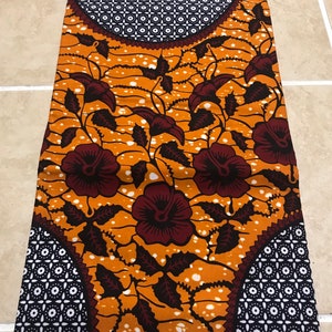 May include: Orange and brown fabric with a floral pattern and two large circles with a geometric design. The fabric has the text "VERITABLE WAX ABC" and "ABRIQUE AU GHANA" printed on it.