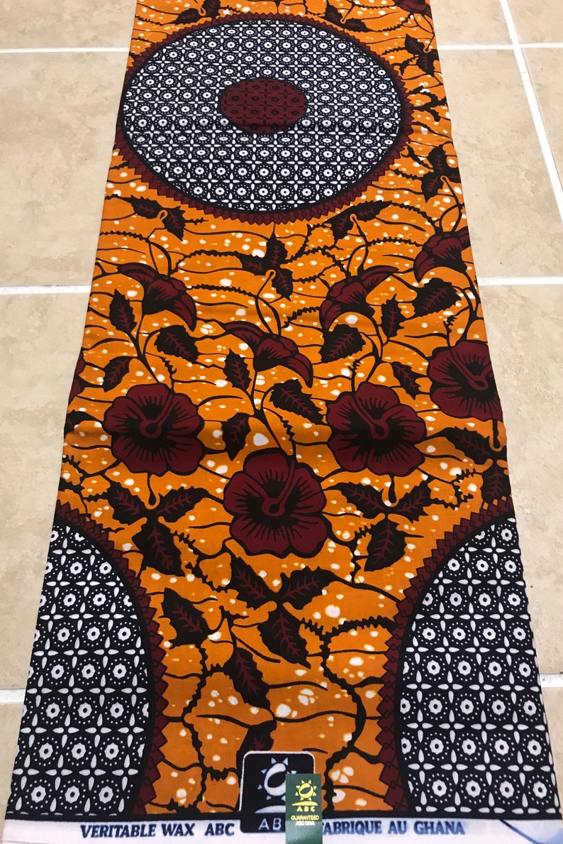 May include: Orange and brown fabric with a floral pattern and two large circles with a geometric design. The fabric has the text "VERITABLE WAX ABC" and "ABRIQUE AU GHANA" printed on it.