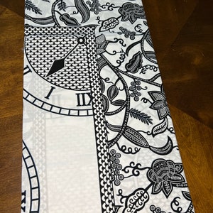 May include: Black and white fabric with a clock face and floral pattern. The fabric is folded in half.