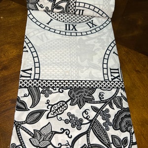 May include: Black and white fabric with a repeating pattern of clocks and floral designs. The fabric has a white background with black clock faces and floral motifs. The fabric is rolled up and the edge of the roll is visible.