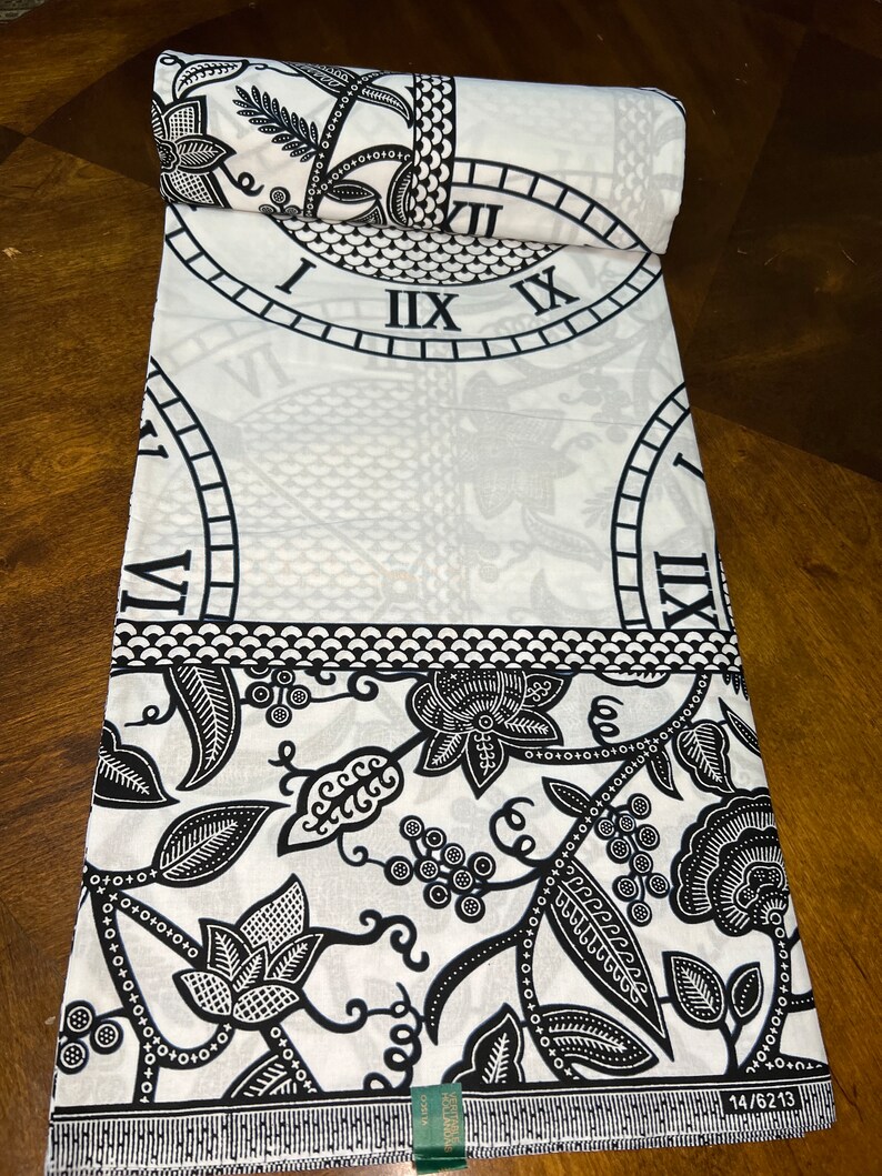 May include: Black and white fabric with a repeating pattern of clocks and floral designs. The fabric has a white background with black clock faces and floral motifs. The fabric is rolled up and the edge of the roll is visible.