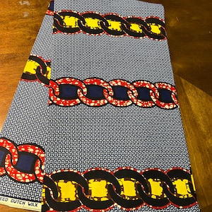 K&ouml;nnte beinhalten: Gefalteter Stoff mit einem sich wiederholenden Kettengliedmuster in Rot, Gelb, Schwarz und Blau. Der Hintergrund ist ein kleines, sich wiederholendes wei&szlig;es und blaues Muster. Der Stoff ist mit "VERITABLE WAX" beschriftet.