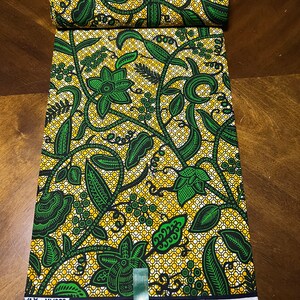 K&ouml;nnte beinhalten: Ein gr&uuml;ner und gelber Stoff mit einem Blumenmuster. Der Stoff ist gefaltet und hat ein Etikett mit der Aufschrift "Wax Vlisco".