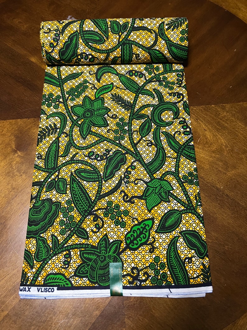 K&ouml;nnte beinhalten: Ein gr&uuml;ner und gelber Stoff mit einem Blumenmuster. Der Stoff ist gefaltet und hat ein Etikett mit der Aufschrift "Wax Vlisco".