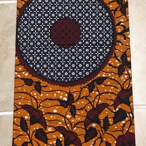 May include: A brown and white fabric with a floral pattern. The fabric has a repeating pattern of brown flowers with white centers and white dots on a brown background. The fabric also has a repeating pattern of black and white squares. The fabric has the text "VERITABLE WAX ABC" and "ABRIQUE AU GHANA" printed on it.