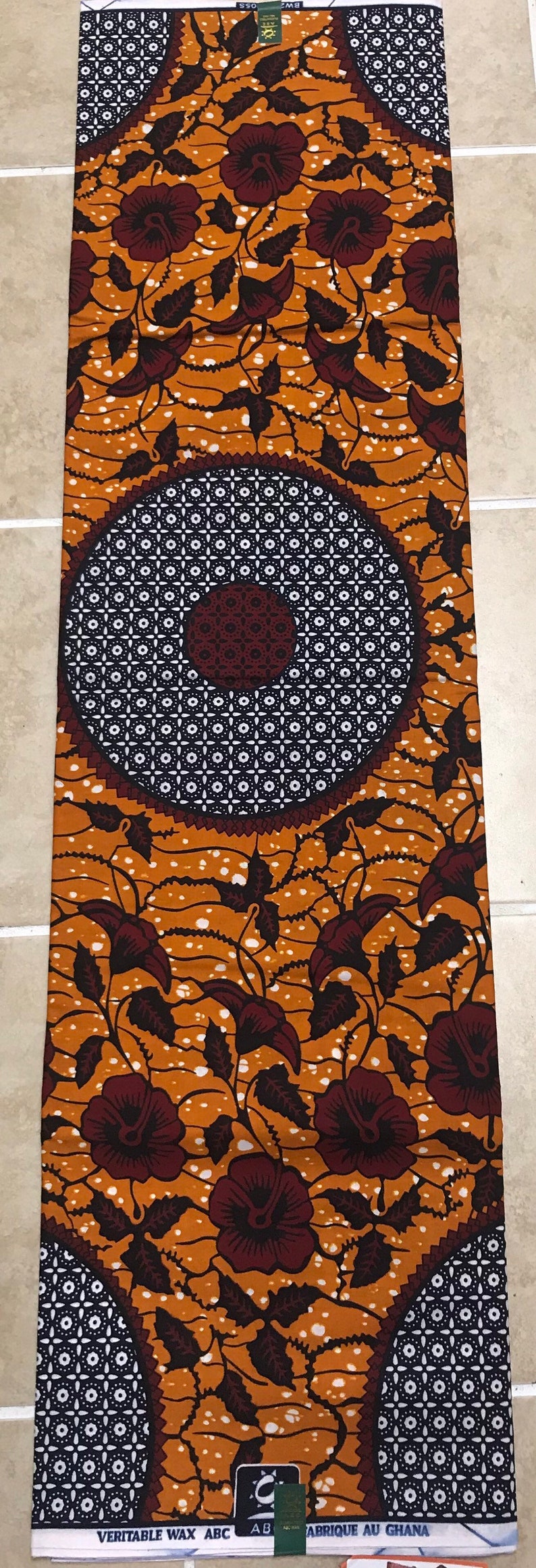 May include: A brown and white fabric with a floral pattern. The fabric has a repeating pattern of brown flowers with white centers and white dots on a brown background. The fabric also has a repeating pattern of black and white squares. The fabric has the text "VERITABLE WAX ABC" and "ABRIQUE AU GHANA" printed on it.