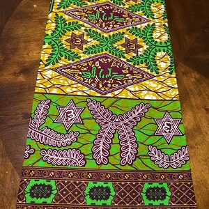 Peut inclure: Un tissu vert, marron et or avec un motif g&eacute;om&eacute;trique compos&eacute; d'&eacute;toiles et de feuilles. Le tissu porte l'inscription "VERITAB AX HOLLANDAIS VLISCO" imprim&eacute;e sur le bord inf&eacute;rieur.