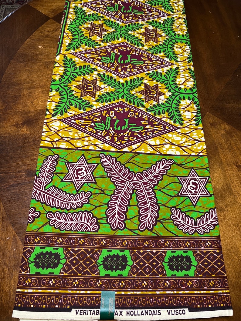 Peut inclure: Un tissu vert, marron et or avec un motif g&eacute;om&eacute;trique compos&eacute; d'&eacute;toiles et de feuilles. Le tissu porte l'inscription "VERITAB AX HOLLANDAIS VLISCO" imprim&eacute;e sur le bord inf&eacute;rieur.