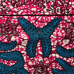 Op de afbeelding: Close-up van een levendige stof met een herhalend patroon. Het ontwerp heeft een magenta achtergrond met witte spikkels en zwarte en blauwe geometrische vormen, waaronder sterren en bladachtige motieven. De stof heeft een getextureerde uitstraling.