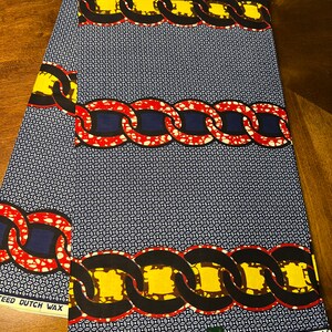 K&ouml;nnte beinhalten: Ein gefaltetes St&uuml;ck Stoff mit Muster. Der Stoff zeigt ein sich wiederholendes Kettenmuster in Gelb, Rot und Schwarz auf einem marineblauen Hintergrund mit einem kleinen wei&szlig;en geometrischen Muster. Der Text "VERITABLE W" ist sichtbar.