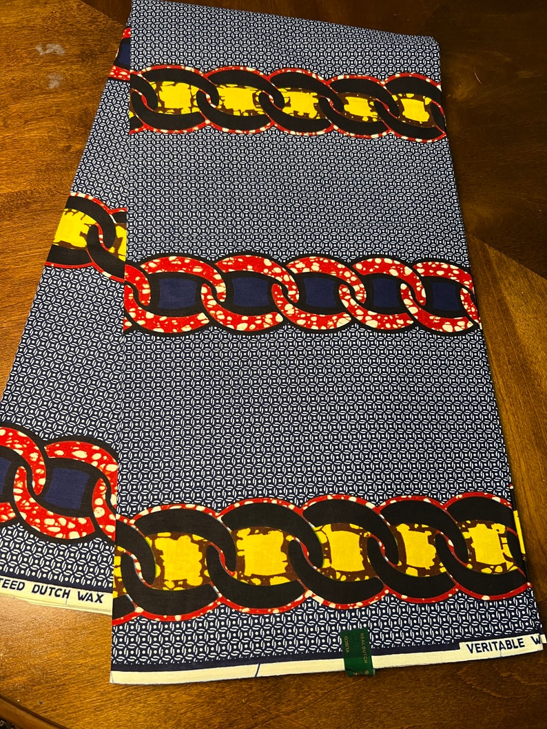 K&ouml;nnte beinhalten: Ein gefaltetes St&uuml;ck Stoff mit Muster. Der Stoff zeigt ein sich wiederholendes Kettenmuster in Gelb, Rot und Schwarz auf einem marineblauen Hintergrund mit einem kleinen wei&szlig;en geometrischen Muster. Der Text "VERITABLE W" ist sichtbar.