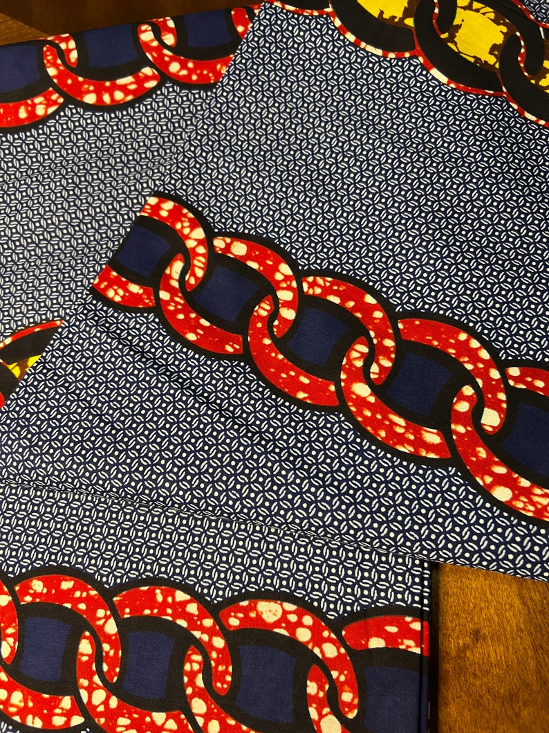 K&ouml;nnte beinhalten: Nahaufnahme eines gemusterten Stoffes mit einem sich wiederholenden Kettendesign in Rot und Wei&szlig; vor einem marineblauen Hintergrund. Der Stoff hat auch ein kleines wei&szlig;es geometrisches Muster. Der Stoff ist gefaltet.