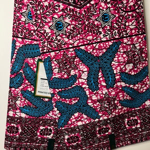 Op de afbeelding: Een gevouwen stof met een levendig patroon. Het ontwerp heeft een roze achtergrond met witte, zwarte en blauwe accenten. De stof heeft een herhalend patroon van sterren, bladeren en geometrische vormen. Een label is zichtbaar met de tekst "PRINTED BY".