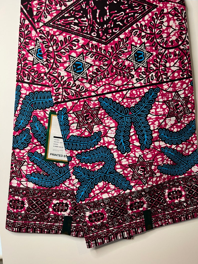 Op de afbeelding: Een gevouwen stof met een levendig patroon. Het ontwerp heeft een roze achtergrond met witte, zwarte en blauwe accenten. De stof heeft een herhalend patroon van sterren, bladeren en geometrische vormen. Een label is zichtbaar met de tekst "PRINTED BY".