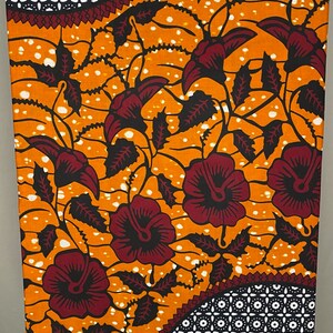 May include: Orange and red African wax print fabric with a floral pattern and geometric designs. The fabric has a white background with small white dots. The fabric is labeled "FABRIQUE AU GHANA" and "ABC".