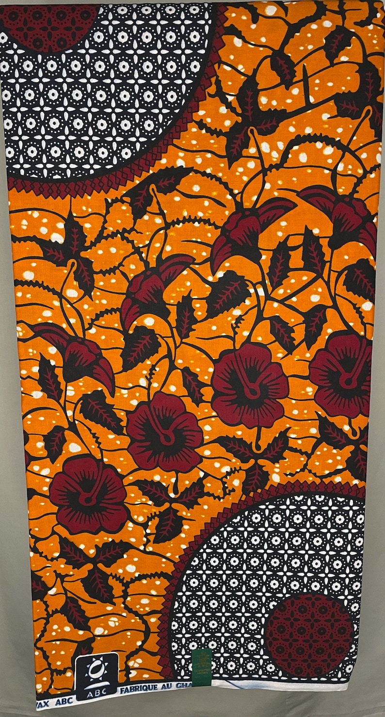 May include: Orange and red African wax print fabric with a floral pattern and geometric designs. The fabric has a white background with small white dots. The fabric is labeled "FABRIQUE AU GHANA" and "ABC".