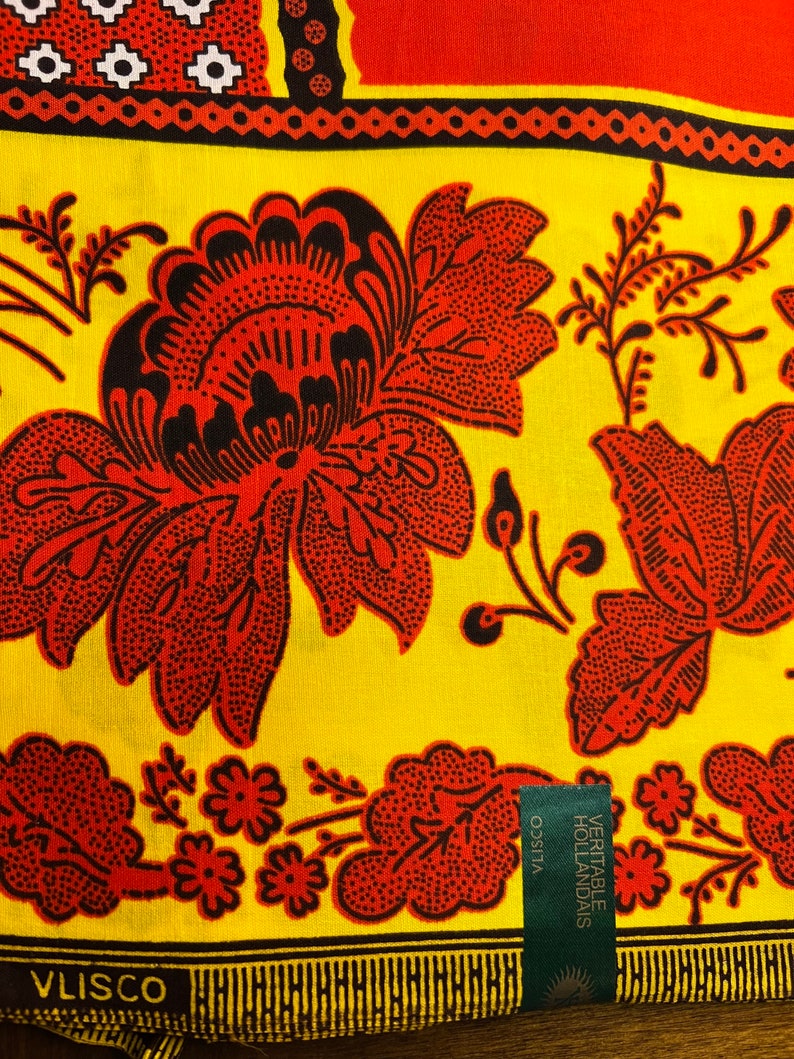 Puede incluir: Una tela amarilla con un patr&oacute;n floral rojo y negro. La tela tiene una etiqueta Vlisco con el texto "VERITABLE HOLLANDAIS".