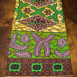 Peut inclure: Tissu vert et marron avec un motif g&eacute;om&eacute;trique compos&eacute; d'&eacute;toiles et de feuilles. Le tissu a un fond blanc avec une bordure marron et le texte "VERITABLE  WAX HOLLANDAIS  VLISCO" imprim&eacute; en bas.