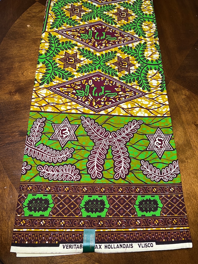 Peut inclure: Tissu vert et marron avec un motif g&eacute;om&eacute;trique compos&eacute; d'&eacute;toiles et de feuilles. Le tissu a un fond blanc avec une bordure marron et le texte "VERITABLE  WAX HOLLANDAIS  VLISCO" imprim&eacute; en bas.