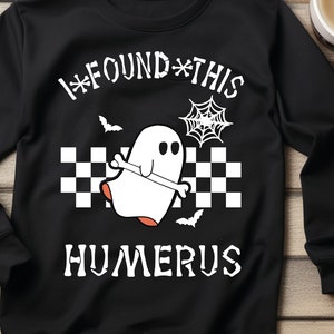 May include: Black sweatshirt with a white ghost design and the text "I*FOUND*THIS HUMERUS" in white. The ghost is wearing a checkered pattern and there are bats and a spider web in the background.
