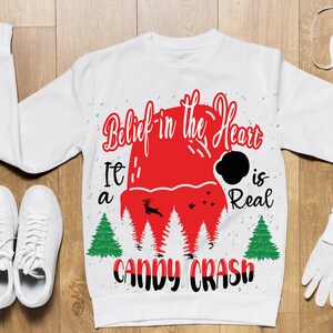 May include: White Christmas sweater with red and black text that reads "Belief in the Heart It's a Candy Crash is Real". The sweater features a red Santa hat with a black pom-pom and a reindeer graphic.