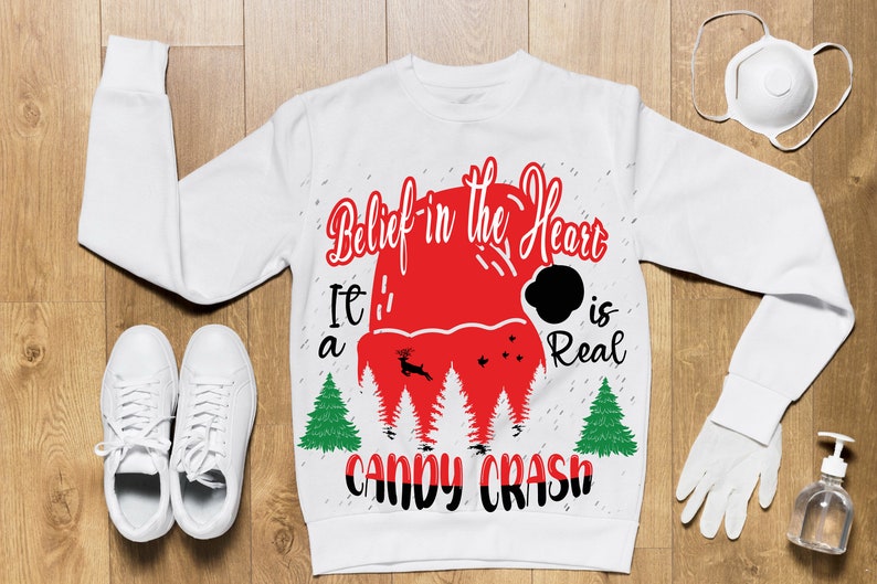 May include: White Christmas sweater with red and black text that reads "Belief in the Heart It's a Candy Crash is Real". The sweater features a red Santa hat with a black pom-pom and a reindeer graphic.