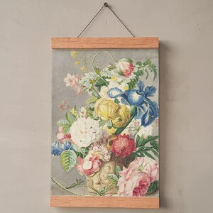 Op de afbeelding: Een bloemenprint ingelijst in licht hout. Het kunstwerk toont een gedetailleerde illustratie van een boeket bloemen in een vaas, met bloemen in roze, geel, blauw en wit. De print wordt opgehangen door een dunne draad.