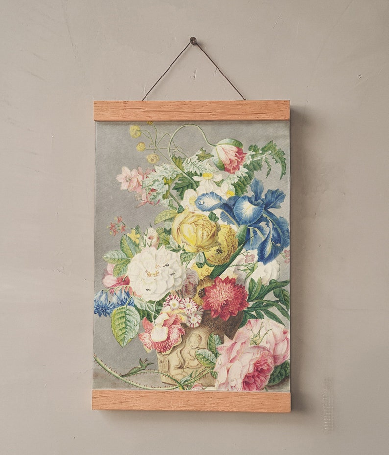Op de afbeelding: Een bloemenprint ingelijst in licht hout. Het kunstwerk toont een gedetailleerde illustratie van een boeket bloemen in een vaas, met bloemen in roze, geel, blauw en wit. De print wordt opgehangen door een dunne draad.