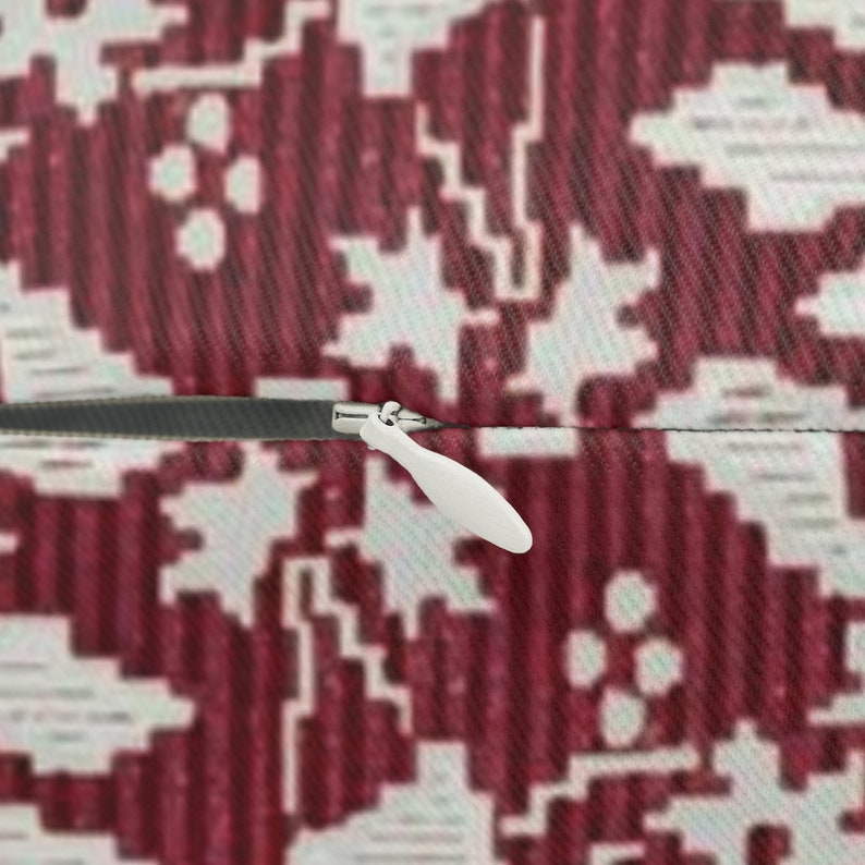 May include: Close-up of a pillow with a burgundy and white geometric pattern. A white zipper pull is visible on the side of the pillow. The pillow has a textured surface.