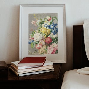Op de afbeelding: Een ingelijste bloemenprint met een boeket kleurrijke bloemen, waaronder rozen en irissen, staat op een donkerhouten nachtkastje. Een stapel boeken met een rode kaft is voor de ingelijste kunst geplaatst. De sc&egrave;ne speelt zich af in een slaapkamer.