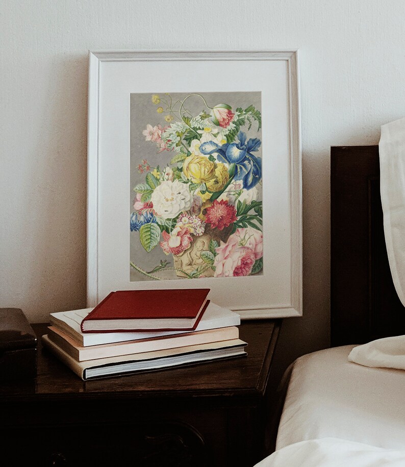 Op de afbeelding: Een ingelijste bloemenprint met een boeket kleurrijke bloemen, waaronder rozen en irissen, staat op een donkerhouten nachtkastje. Een stapel boeken met een rode kaft is voor de ingelijste kunst geplaatst. De sc&egrave;ne speelt zich af in een slaapkamer.