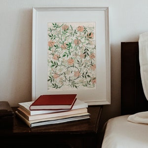 May include: A framed print of a botanical illustration featuring a white and green floral pattern with pink flowers. The print is on a white mat and is displayed on a wooden nightstand with a stack of books.