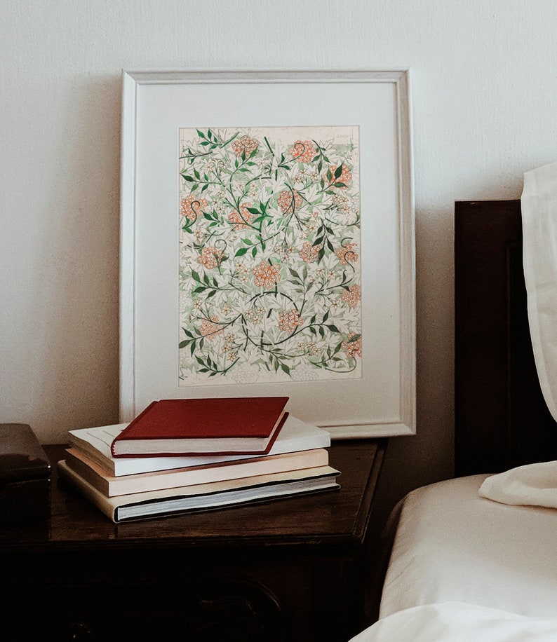 May include: A framed print of a botanical illustration featuring a white and green floral pattern with pink flowers. The print is on a white mat and is displayed on a wooden nightstand with a stack of books.