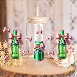 Op de afbeelding: Een heldere glazen beker met een bamboe deksel en rietje, geflankeerd door twee groene Sprite-flessen en een Sprite-blikje, allemaal versierd met roze strikken. De items zijn gerangschikt op een houten schijf, met decoratieve elementen.