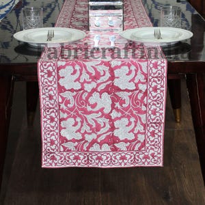 May include: A red and white floral patterned table runner. The runner features a detailed white floral design on a red background. It is rectangular and placed on a dark wooden table. Place settings with plates and cutlery are visible.