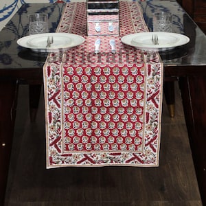 May include: A red table runner with a repeating floral pattern in white and gold. The runner is placed on a dark wooden table, with white plates, silverware, and glasses visible. The runner is approximately 152 cm long.