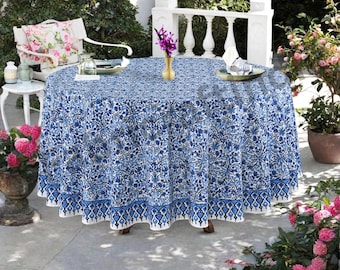 May include: A round tablecloth with a blue and white floral pattern. The tablecloth is set on a wooden table outdoors, with a white chair and flowers in the background. The tablecloth has a decorative border.