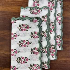 May include: A stack of four white cloth napkins with a floral pattern in pink and green. Each napkin has a scalloped edge in green. The napkins are arranged on a wooden surface.