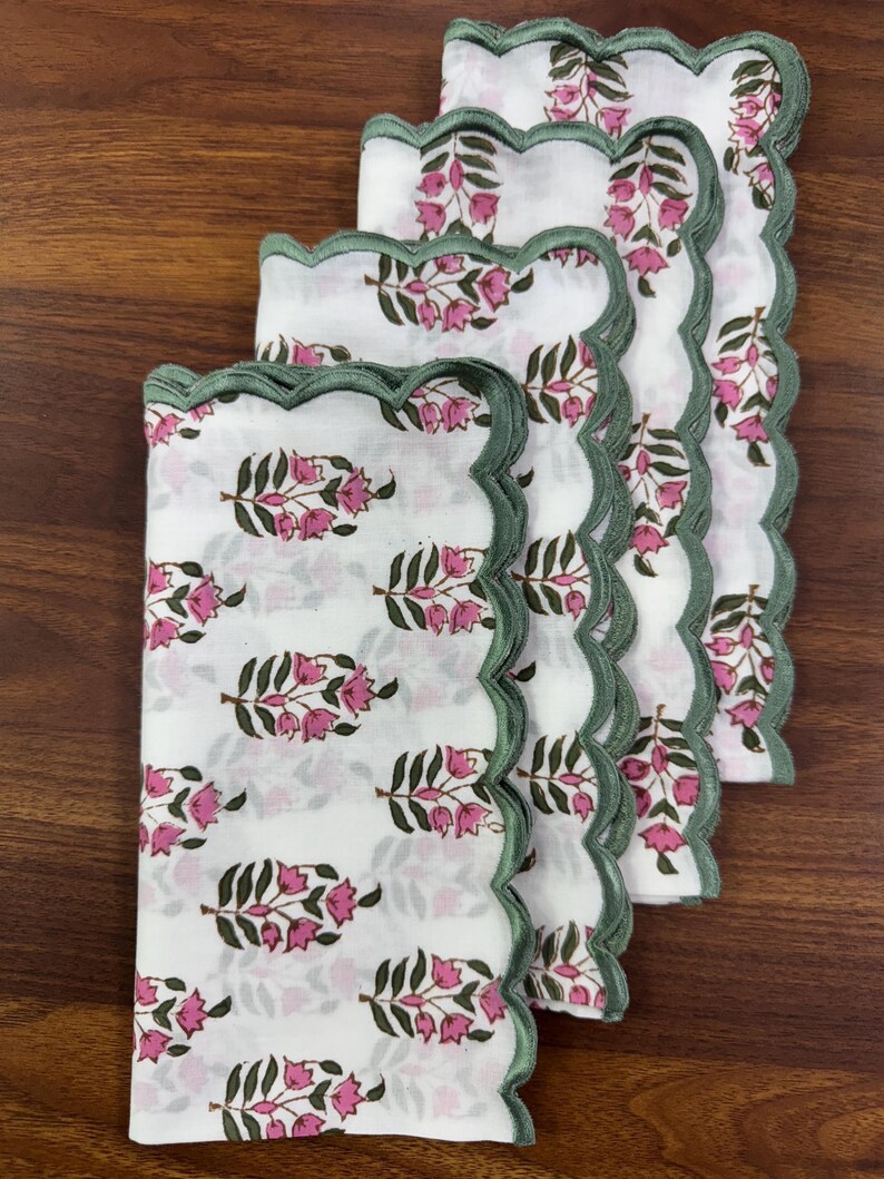 May include: A stack of four white cloth napkins with a floral pattern in pink and green. Each napkin has a scalloped edge in green. The napkins are arranged on a wooden surface.